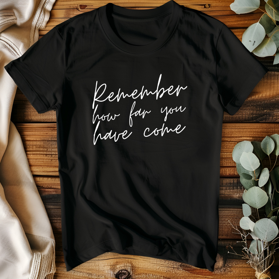Black t-shirt with 'Remember how far you have come' text on a wooden surface with plants.