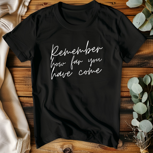 Black t-shirt with 'Remember how far you have come' text on a wooden surface with plants.
